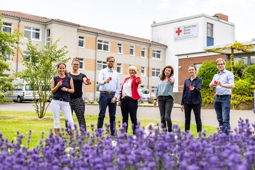 DRK Mitarbeitende stehen mit einem Roten Kreuz in der Hand vor dem DRK-Standort Raisdorf