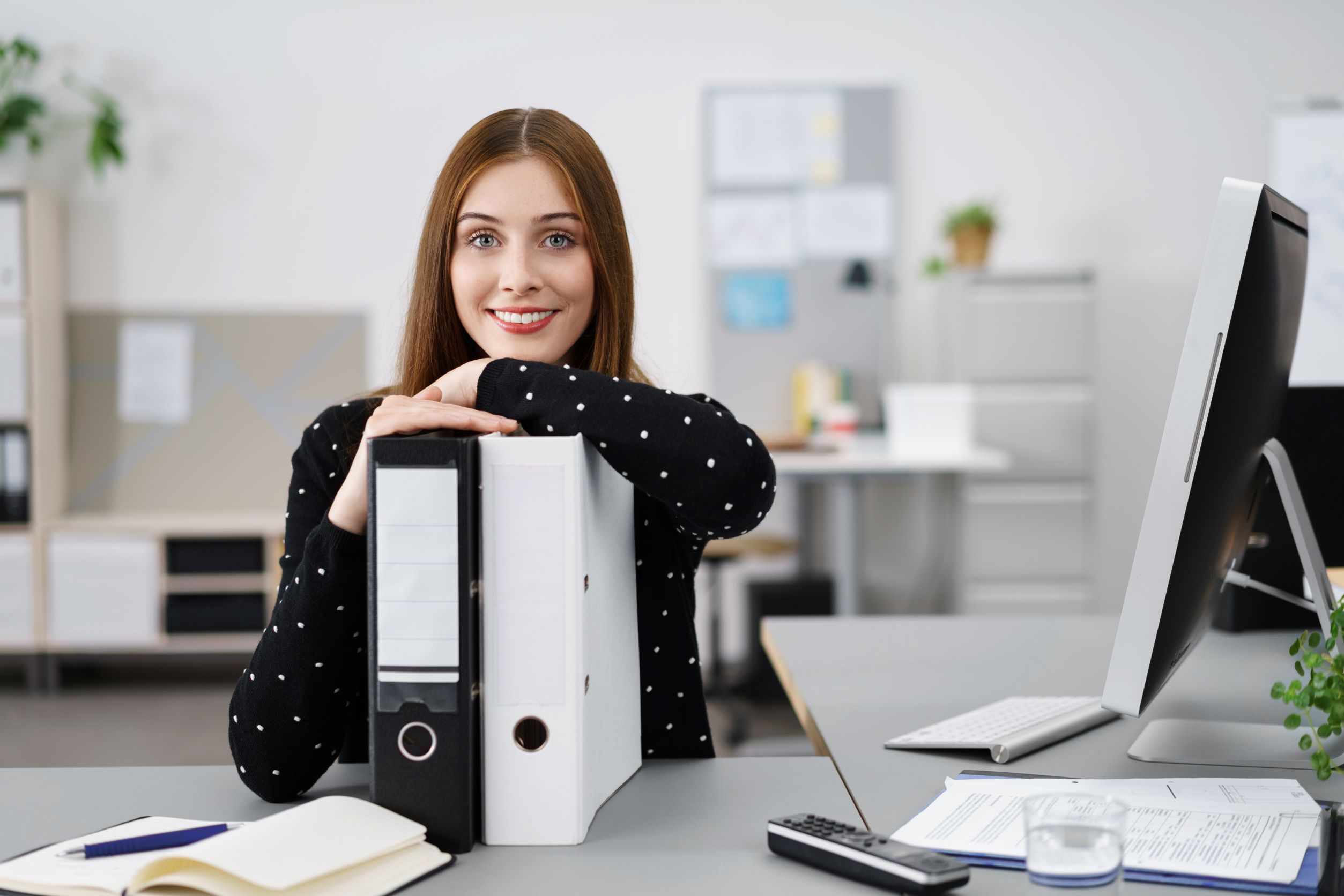Bürokauffrau stützt sich am Schreibtisch auf zwei Ordnern ab und lächelt